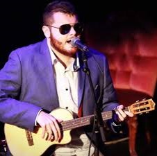 Connor is wearing a white shirt & purple jacket, signature black Ray Ban sunglasses, holding his acoustic guitar against a black stage background.