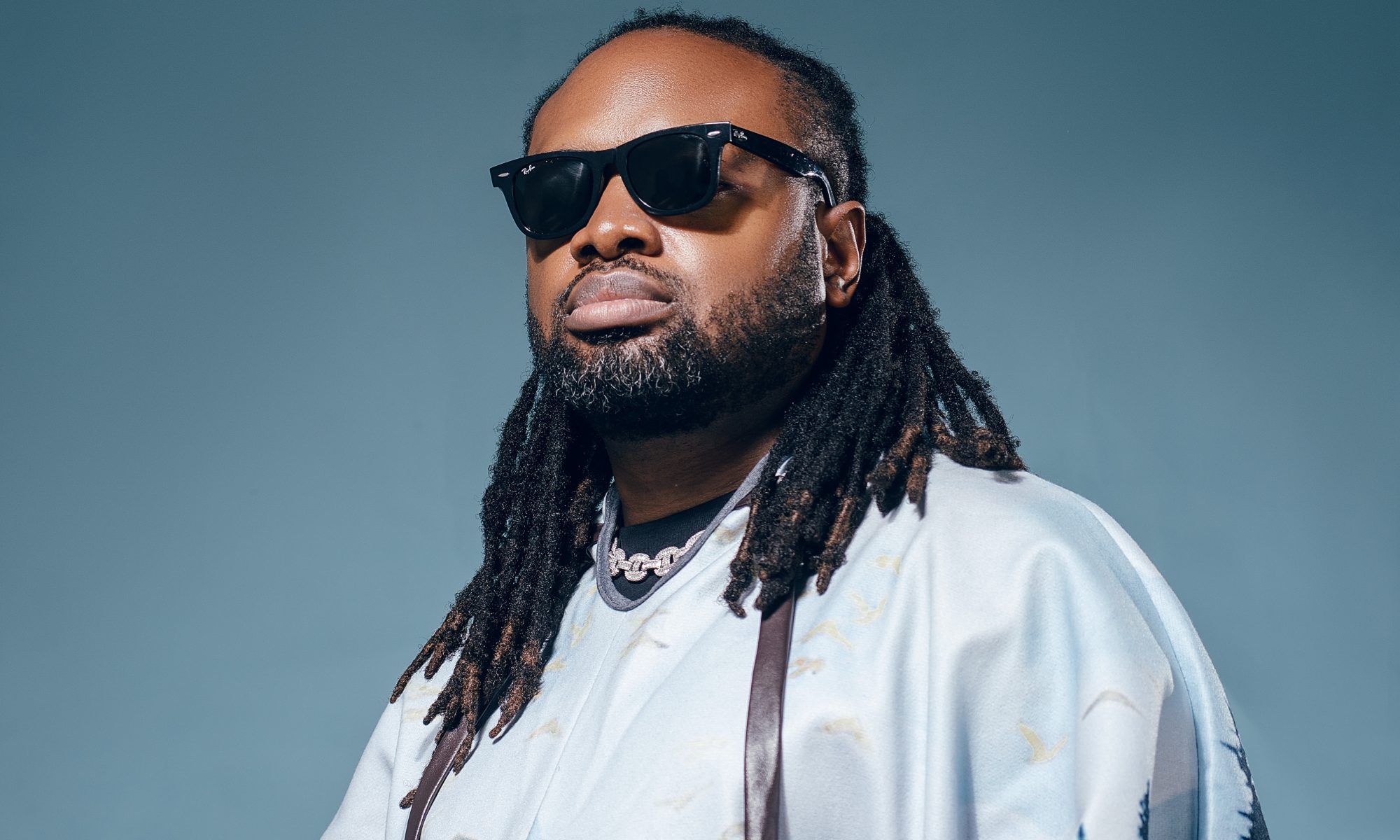 Photo of Cobhams Asuquo in a blue short sleeve shirts with sunglasses against a blue turquoise background.