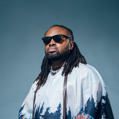 Photo of Cobhams Asuquo in a blue short sleeve shirts with sunglasses against a blue turquoise background.
