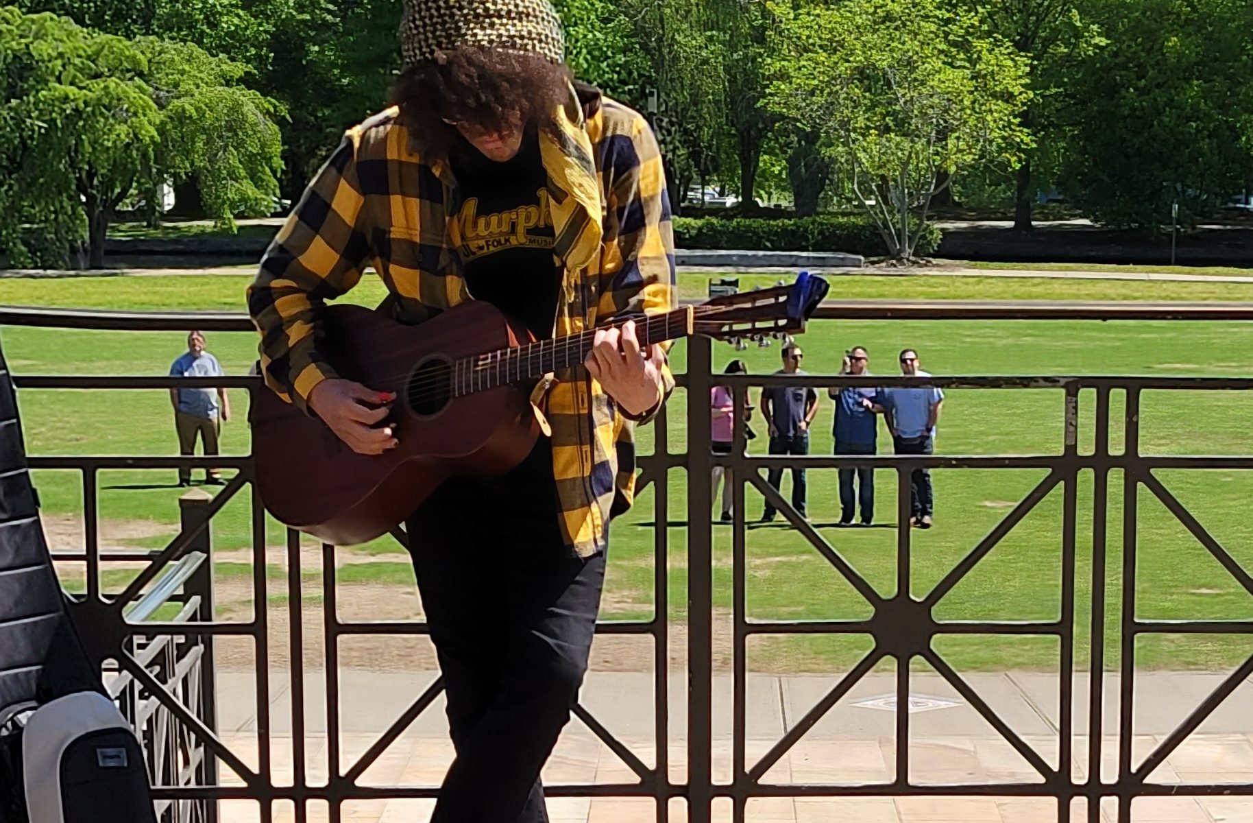 photo of Murphy, wearing a horizontal stripe brown and yellow wind sheeter with a brown beanie, playing acoustic guitar, leaning against a metal grey fence / handrail, looking out to green grass and trees with a blue sky in the background.