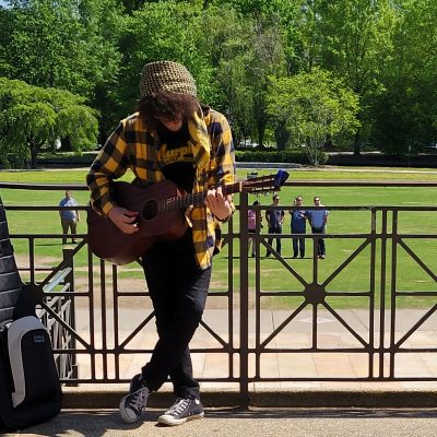 photo of Murphy, wearing a horizontal stripe brown and yellow wind sheeter with a brown beanie, playing acoustic guitar, leaning against a metal grey fence / handrail, looking out to green grass and trees with a blue sky in the background.
