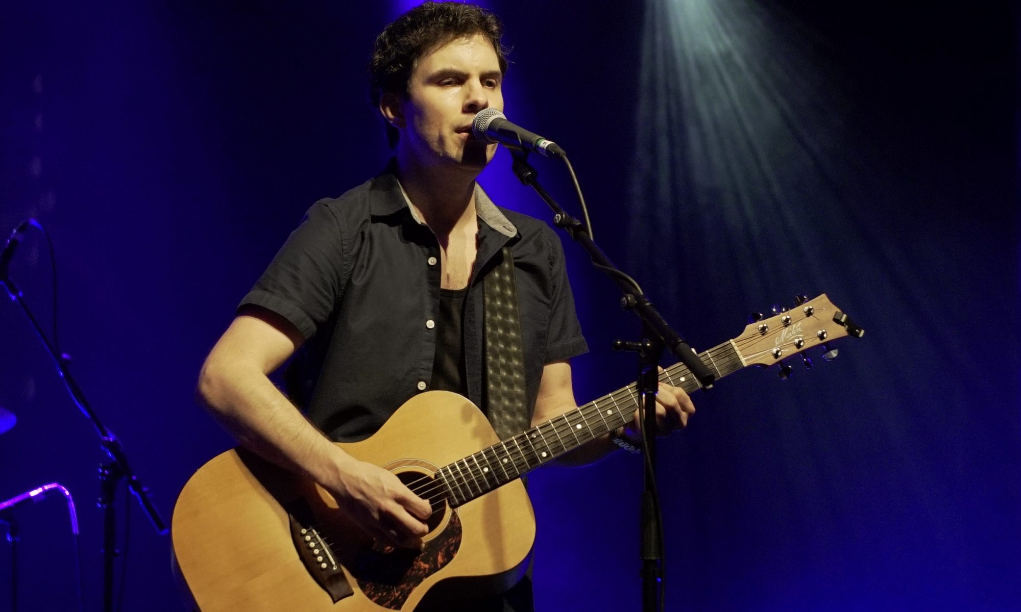 Robert standing on stage with his guitar in a dark grey short sleeve jacket and a dark blue stage background under stage lights.