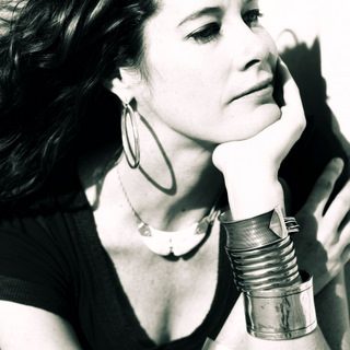 black-and-white Photo of Caitlin Smith sitting on a chair, wearing a black sleeveless dress with long, black hair, with her chin resting on her left hand propped up on her elbow.