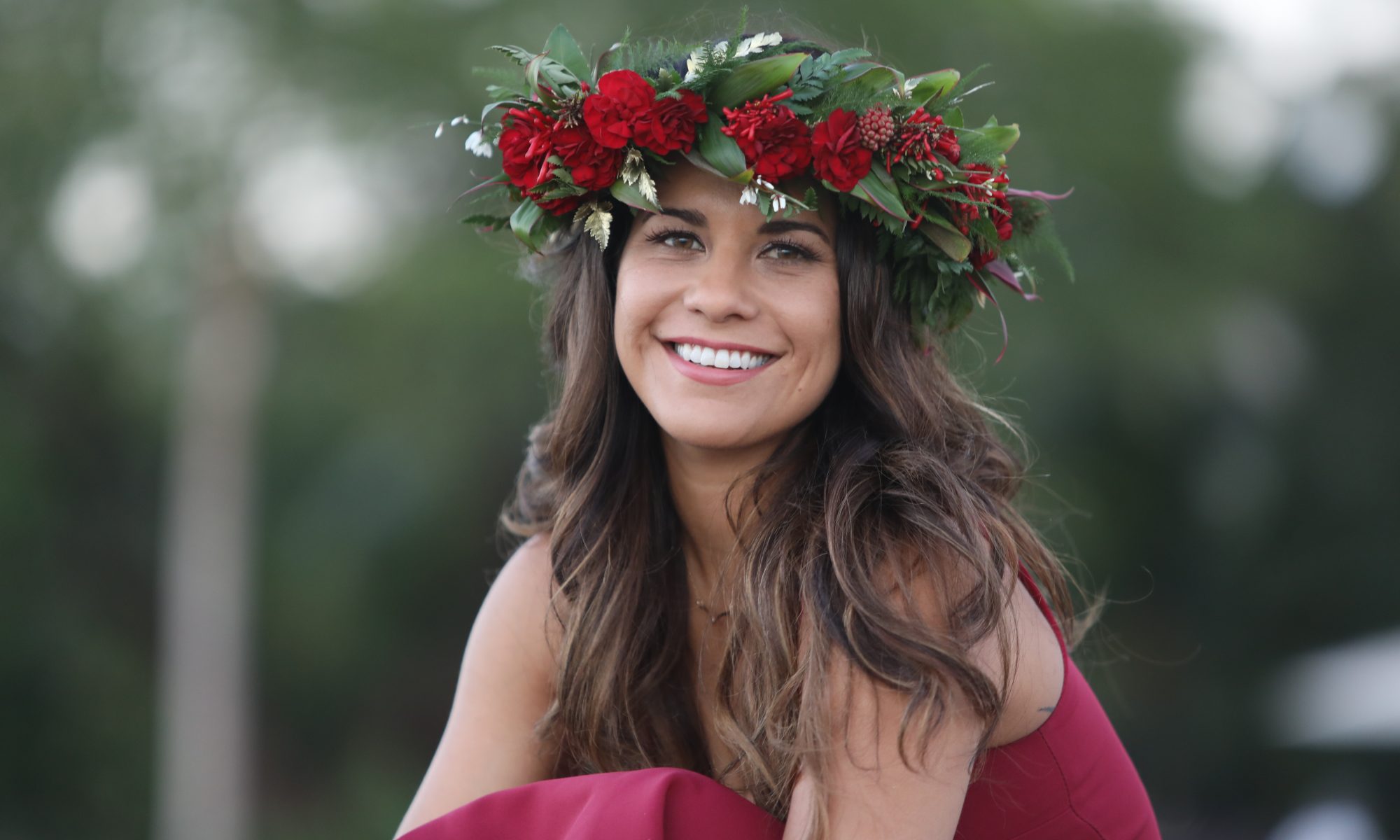Picture of Kimie Miner in a beautiful maroon dress wearing a dark pink rose headdress with long brown hair, smiling, against a green tree background that is out of focus.