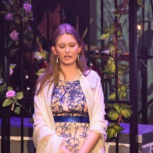 Kirsten pictured in a long cream and colourful full-length dress and jacket, ready to perform against a dark background with purple roses.