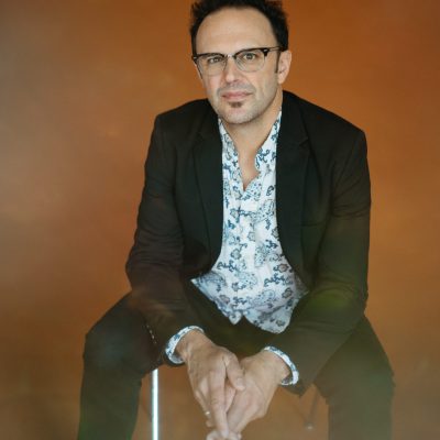 Photo of Mark Erelli wearing glasses, a blue and white colourful shirt beneath a black jacket and pants, sitting on a chair with hands clasped, with arms resting on his knees against an orange background.